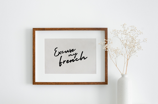 Excuse my French art print in a monochrome colour scheme. The typography is black, in a handwritten style against an off white/greige background. The print is displayed in a wall hung wooden frame, next to a white vase with foliage.