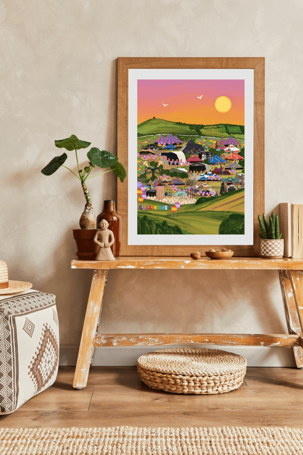 This is a mock up of a Glastonbury Festival print in a frame. Featuring all stages in a view of Glastonbury Festival featuring all areas of the festival including the iconic Pyramid stage, Ribbon Tower and Arcadia, also featuring the Glastonbury Tor against a pink sky.