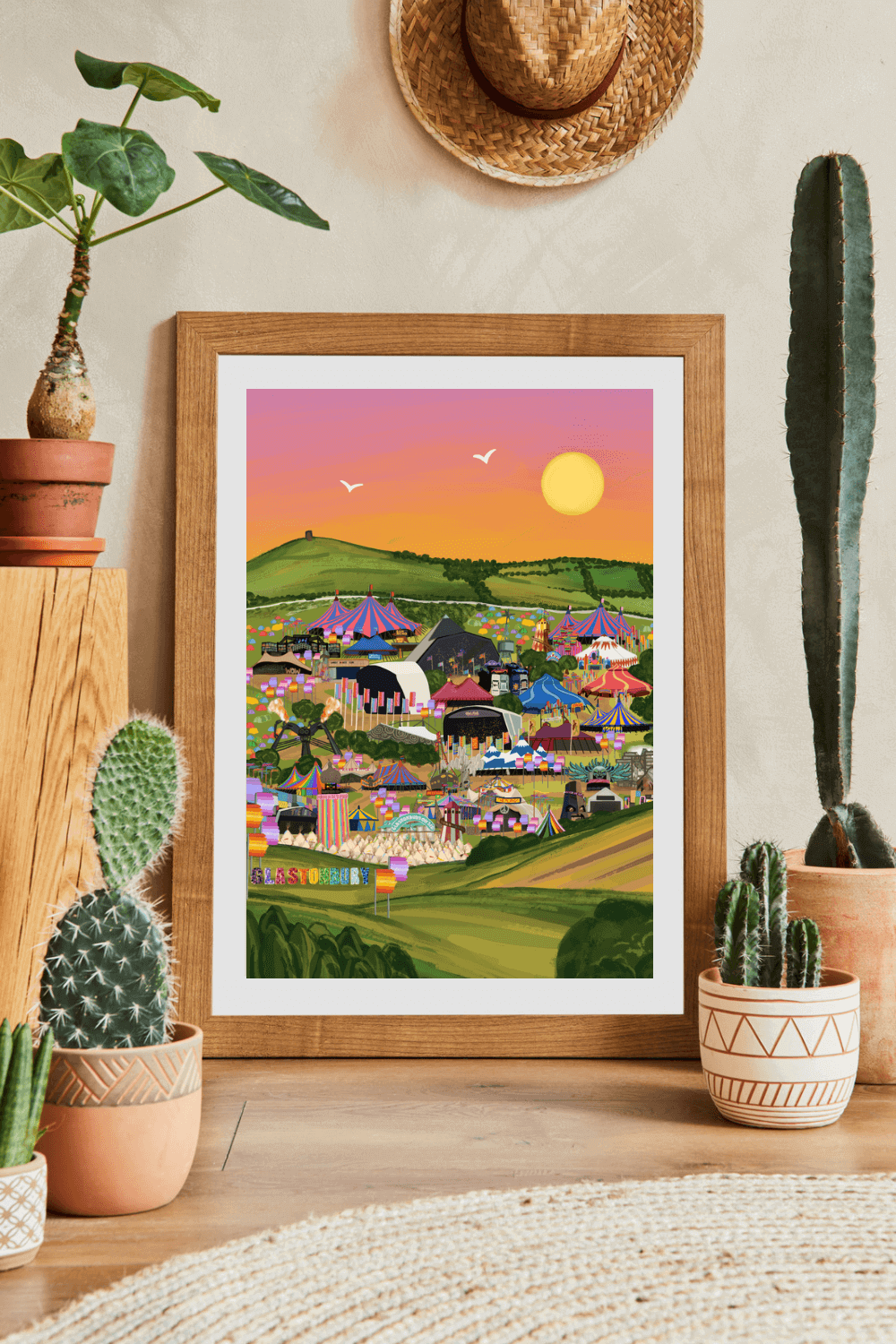 This is an art print of Glastonbury Festival. It depicts the festival stages, flags, Glastonbury sign using vibrant colours. A view of Glastonbury Festival featuring all areas of the festival including the iconic Pyramid stage, Ribbon Tower and Arcadia, also featuring the Glastonbury Tor against a pink sky.