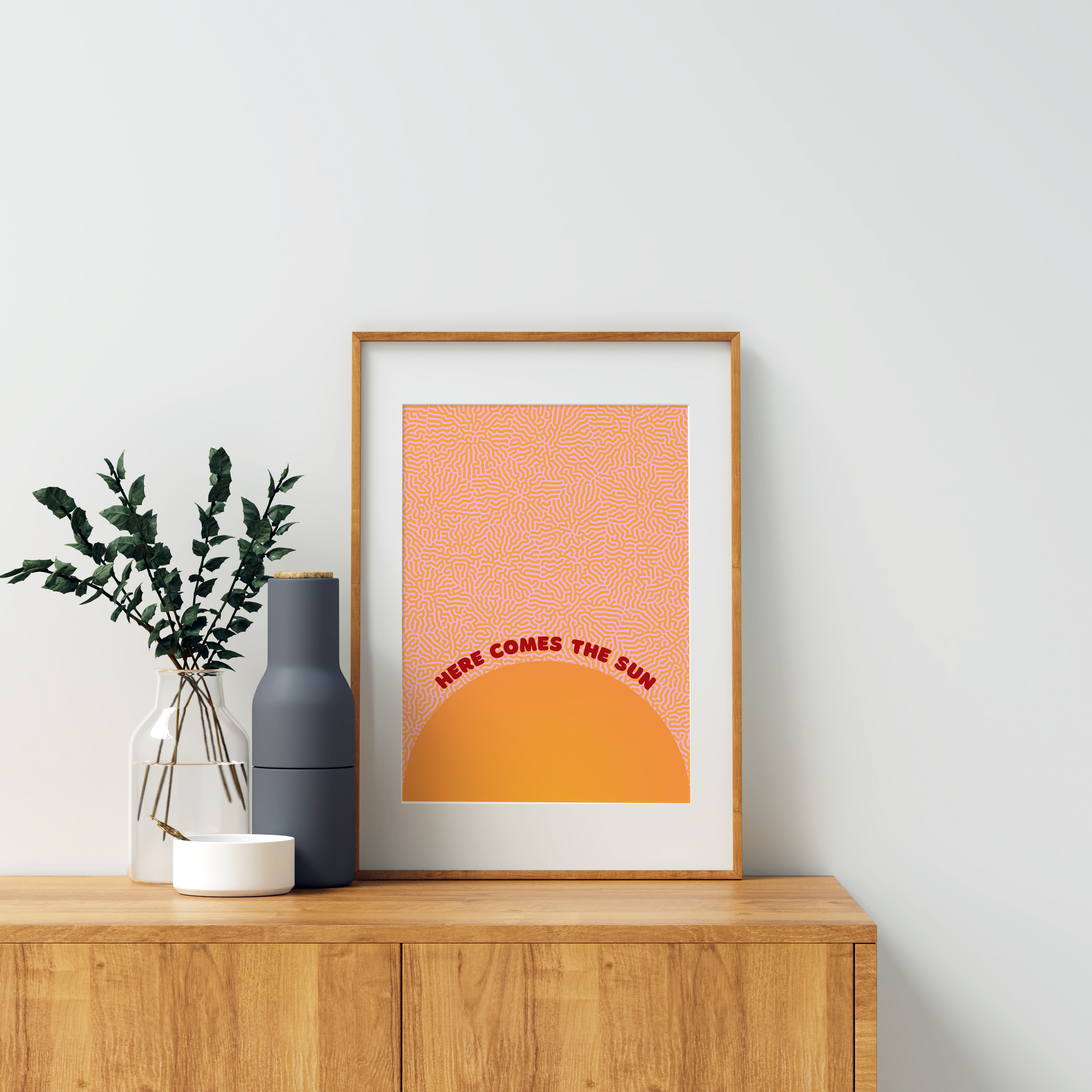 Here comes the sun art print, in orange and red colour scheme. Print is framed in a wooden frame, on a sideboard with indoor plants, foliage and vase. For display purposes only.