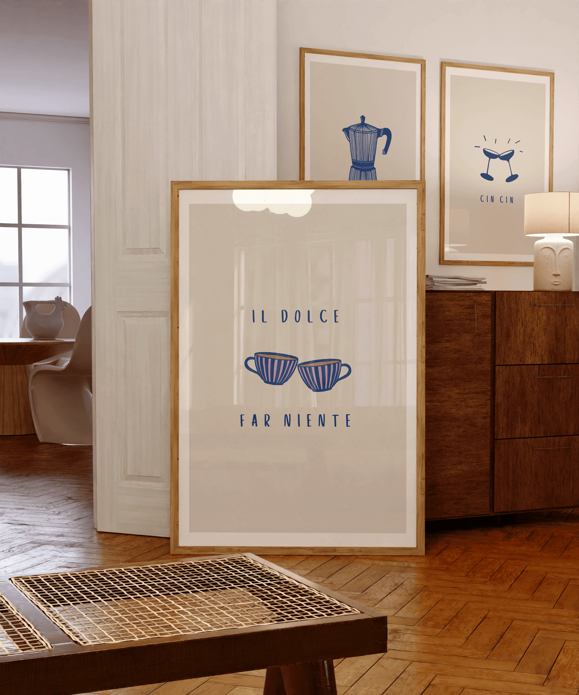 An illustration of two cups of tea or coffee, in navy and pink stripes, against a neutral cream-beige background with the typography "il dolce far niente". Italian print is framed leaning against a sideboard with two framed wall hung prints in background.