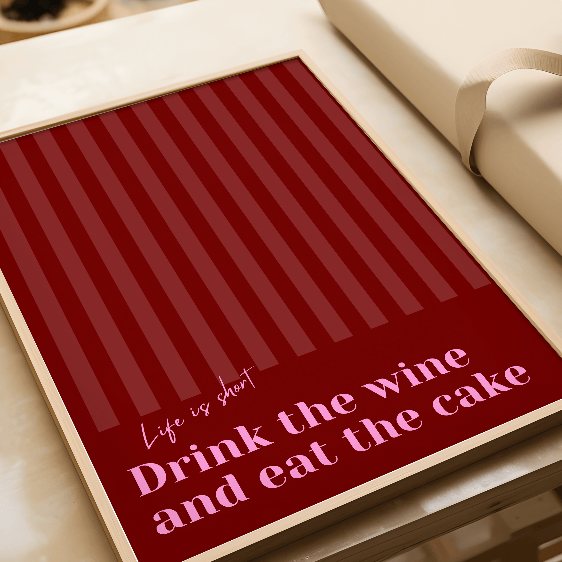 Red and pink typography print, in a wooden frame laying on a table featuring the quote "Life is short. Drink the wine and eat the cake"